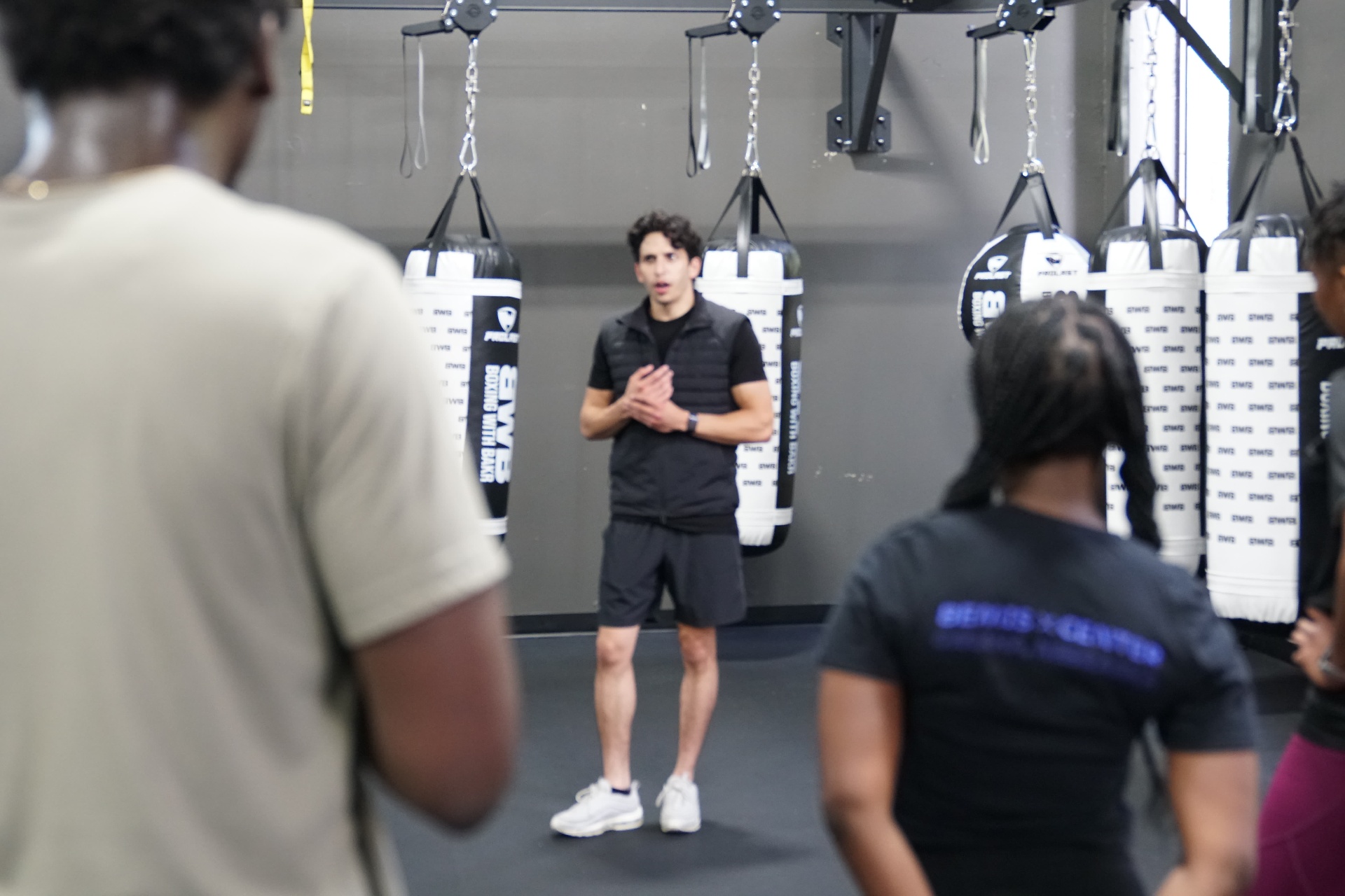 Group Classes — Coach Bakr addressing the class between the heavy bags, students listening in the foreground