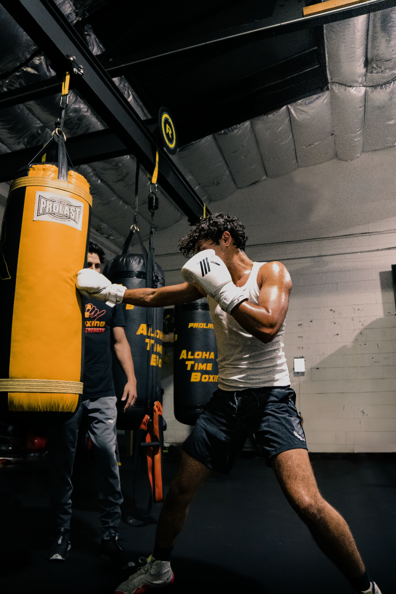 Raj on the heavy bag, white tank, mid-cross at the gym